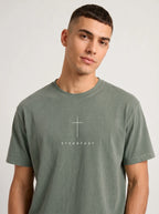 Person wearing a faded sage green T-shirt with a white cross above the text “Steadfast.”