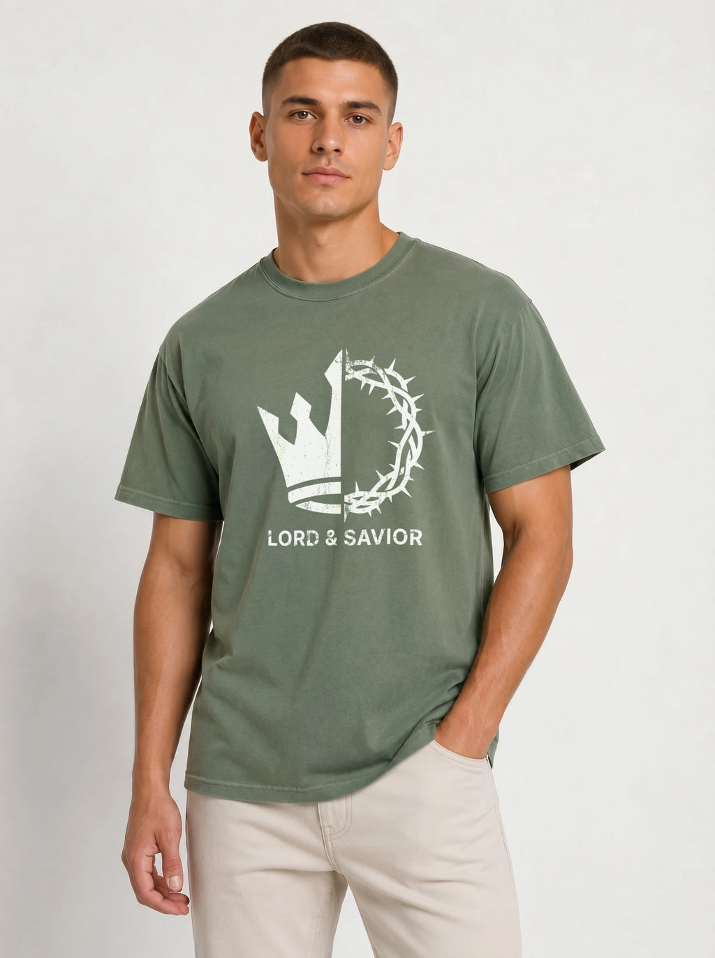 A man wearing an olive green t-shirt with a white graphic of a crown of thorns and the text 'LORD & SAVIOR' on the front, paired with light beige pants, against a plain light gray background.