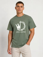 A man wearing an olive green t-shirt with a white graphic of a crown of thorns and the text 'LORD & SAVIOR' on the front, paired with light beige pants, against a plain light gray background.