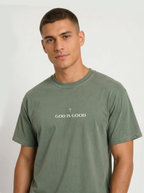 Person wearing a muted green T-shirt with a small cross above the text “God Is Good.”