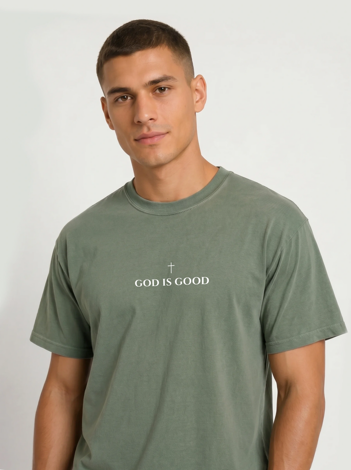 Person wearing a muted green T-shirt with a small cross above the text “God Is Good.”