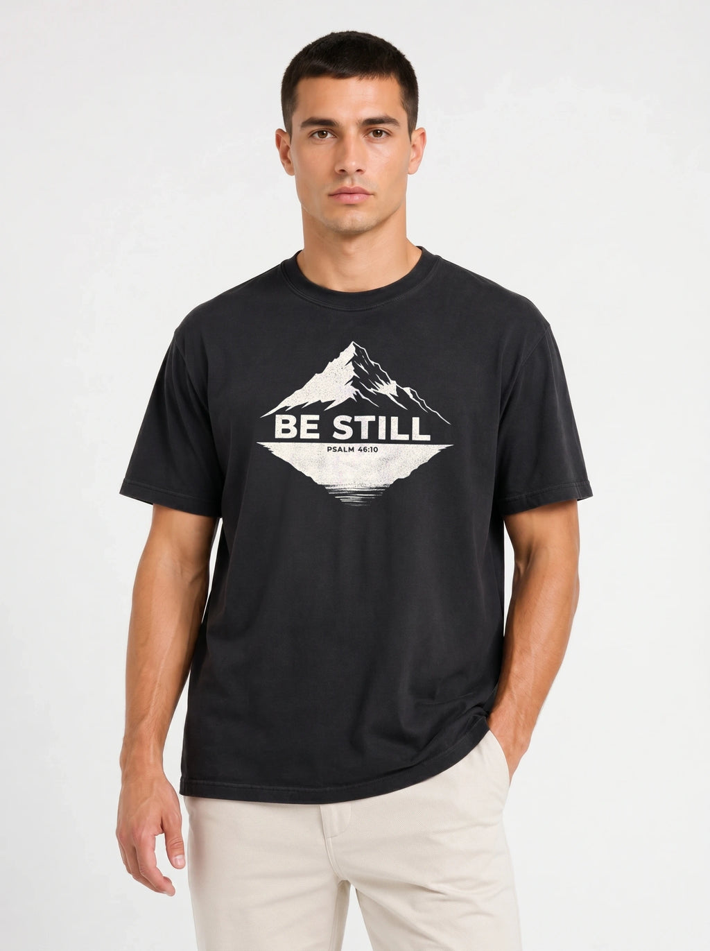 A man wearing a black crewneck t-shirt with a white mountain graphic and the text 'BE STILL' on the chest, paired with light beige pants, against a plain white studio background.