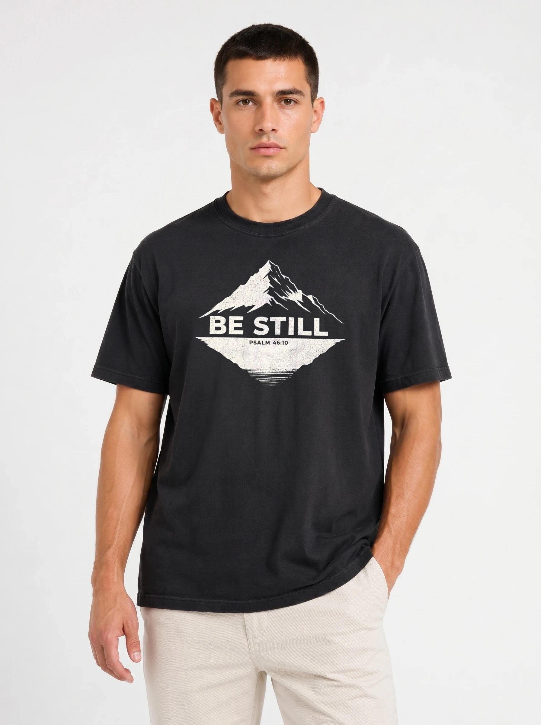 A man wearing a black crewneck t-shirt with a white mountain graphic and the text 'BE STILL' on the chest, paired with light beige pants, against a plain white studio background.