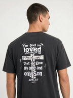 A man wearing a dark charcoal gray t-shirt, viewed from the back. The shirt features a distressed white graphic with the text 'For God so loved the world that He gave His one and only Son JOHN 3:16' surrounding a cracked cross design.