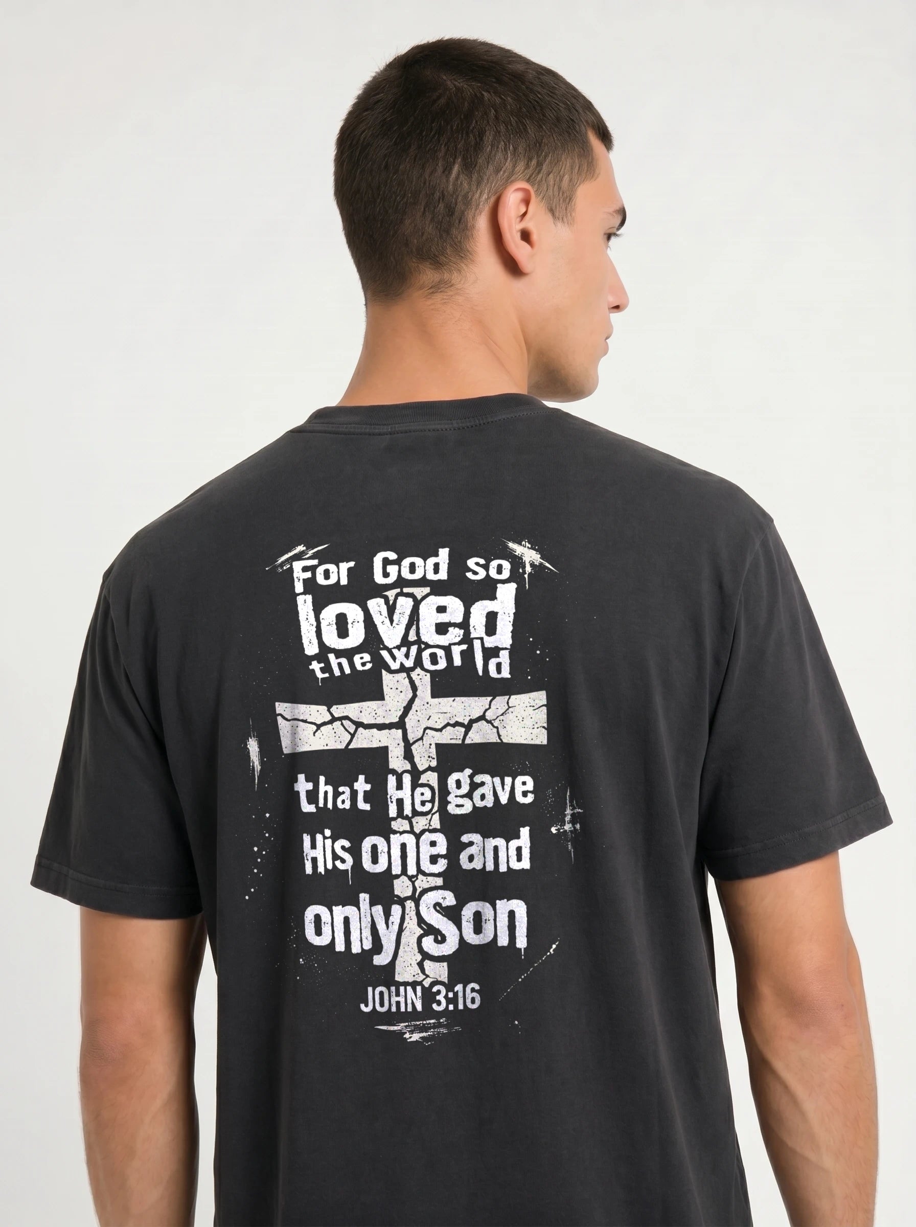 A man wearing a dark charcoal gray t-shirt, viewed from the back. The shirt features a distressed white graphic with the text 'For God so loved the world that He gave His one and only Son JOHN 3:16' surrounding a cracked cross design.