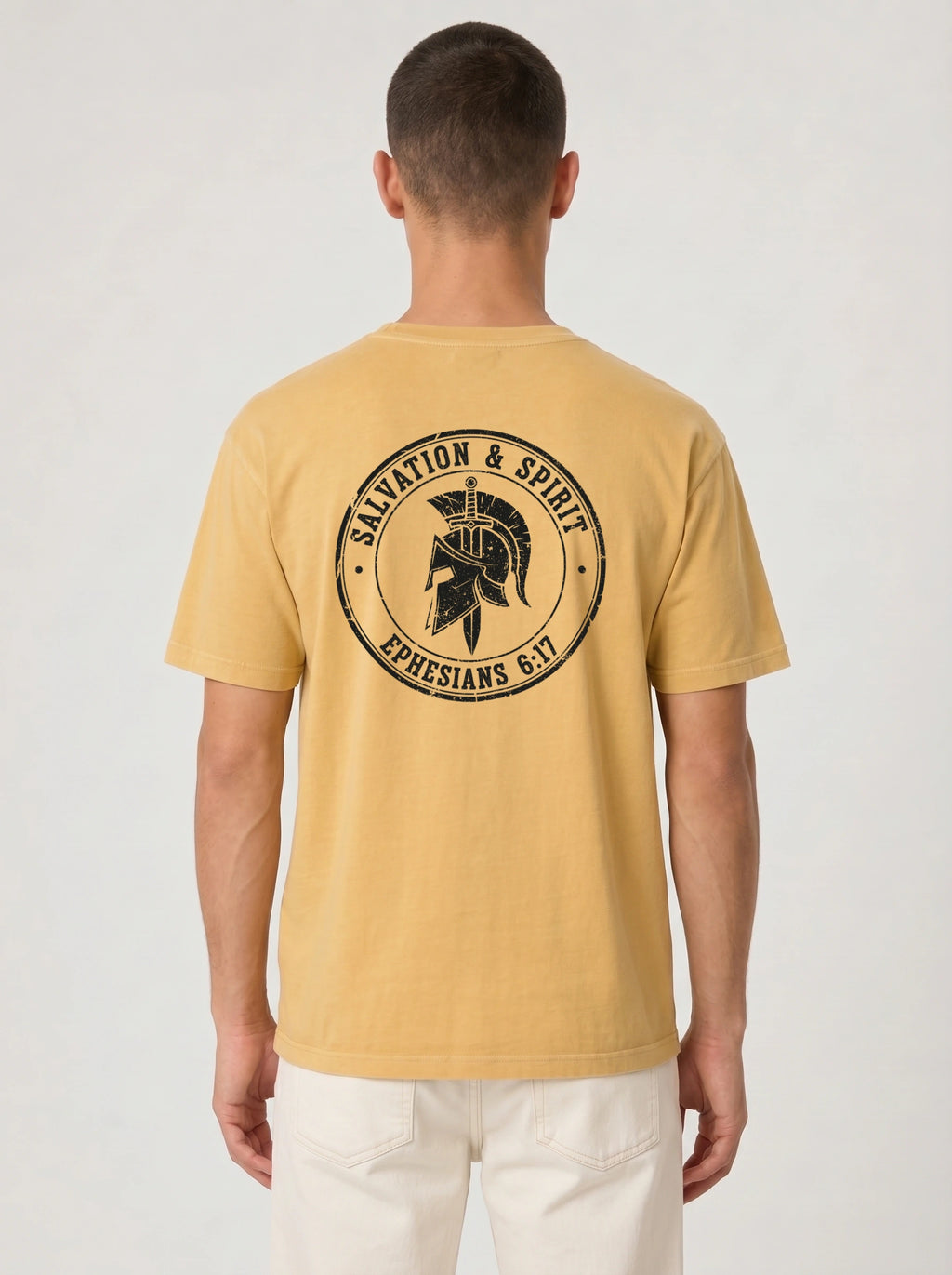 A man viewed from behind wearing a mustard yellow t-shirt with a black circular graphic on the back featuring a Spartan helmet and the text 'SALVATION & SPIRIT EPHESIANS 6:17'.