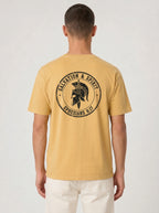 A man viewed from behind wearing a mustard yellow t-shirt with a black circular graphic on the back featuring a Spartan helmet and the text 'SALVATION & SPIRIT EPHESIANS 6:17'.