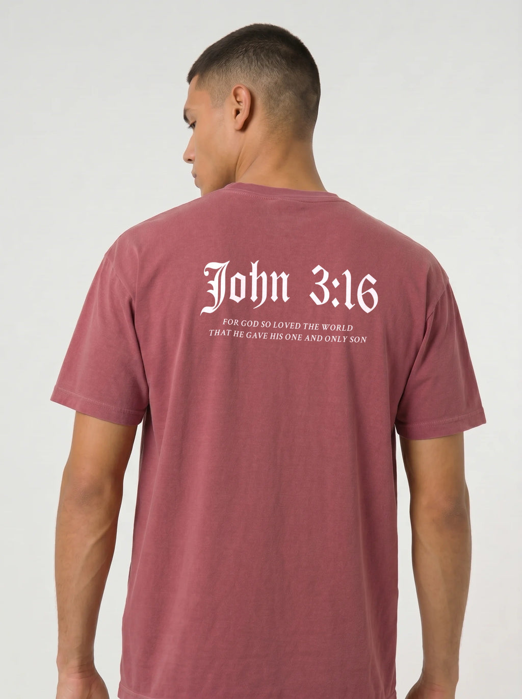A man wearing a dusty rose short-sleeve t-shirt, viewed from the back. The shirt features a 'John 3:16' printed in white Gothic font on the back.