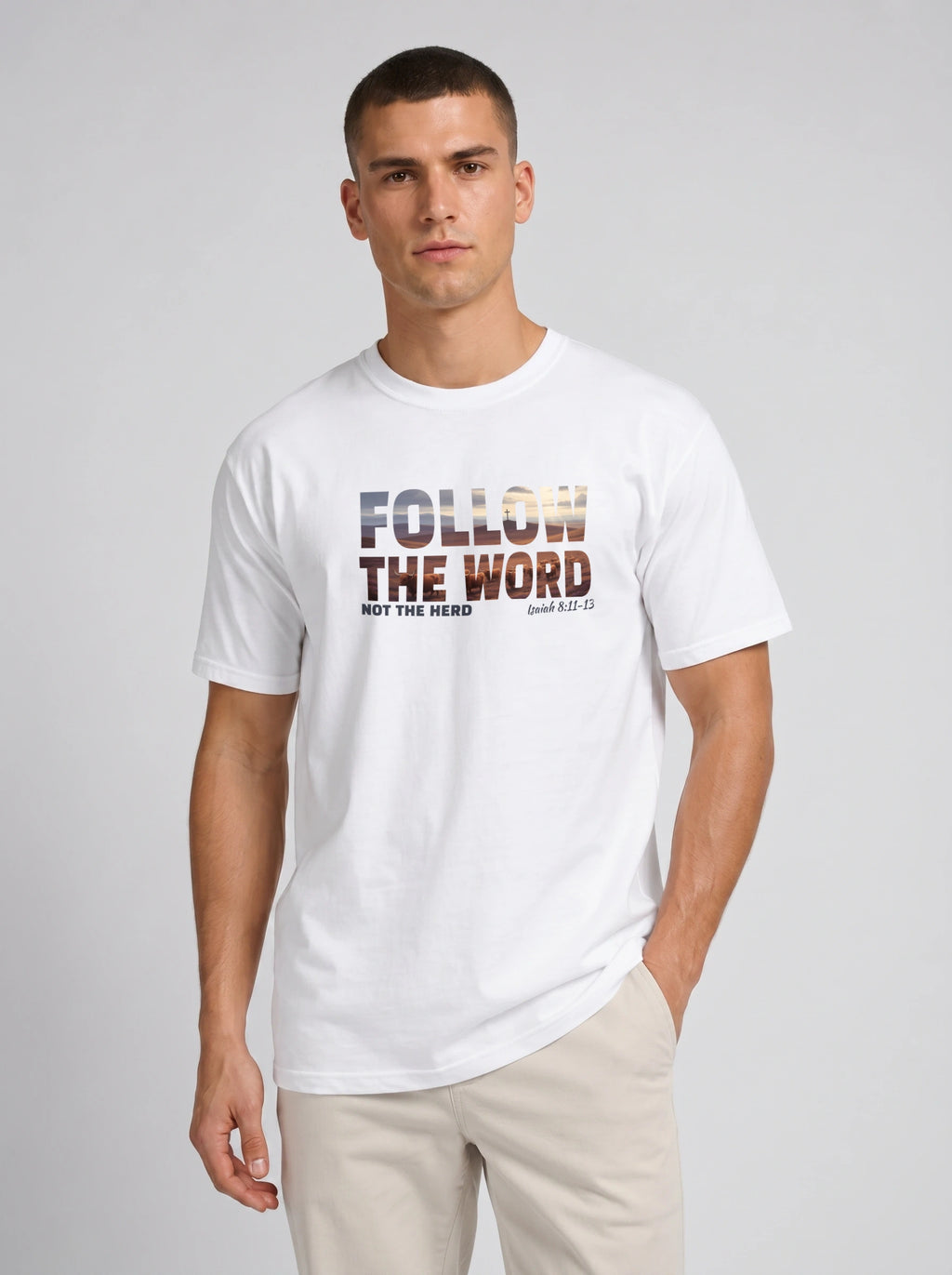 A man wearing a white crew-neck t-shirt with a graphic print that reads 'FOLLOW THE WORD NOT THE HERD' and 'Isaiah 8:11-13' in a textured, multi-toned font. He is also wearing beige trousers, standing against a neutral light gray studio background.