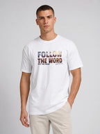 A man wearing a white crew-neck t-shirt with a graphic print that reads 'FOLLOW THE WORD NOT THE HERD' and 'Isaiah 8:11-13' in a textured, multi-toned font. He is also wearing beige trousers, standing against a neutral light gray studio background.