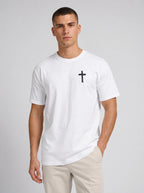 A man wearing a white short-sleeve crewneck t-shirt with a small black cross printed on the left chest, paired with beige trousers, against a light gray studio background.
