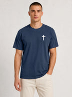 A man wearing a navy blue crewneck t-shirt with a small white cross on the left chest, paired with light beige pants, standing against a light gray studio background.