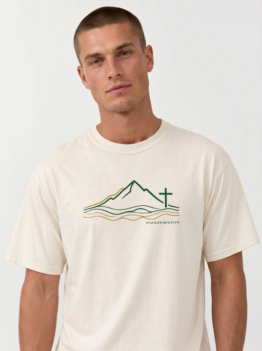 A man wearing a cream-colored t-shirt with a minimalist line art logo featuring a mountain range above flowing river lines, with a Christian cross standing on the right side. The design is drawn in green and gold on a light background, with geographic coordinates printed beneath the landscape.