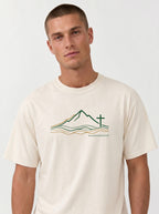 A man wearing a cream-colored t-shirt with a minimalist line art logo featuring a mountain range above flowing river lines, with a Christian cross standing on the right side. The design is drawn in green and gold on a light background, with geographic coordinates printed beneath the landscape.