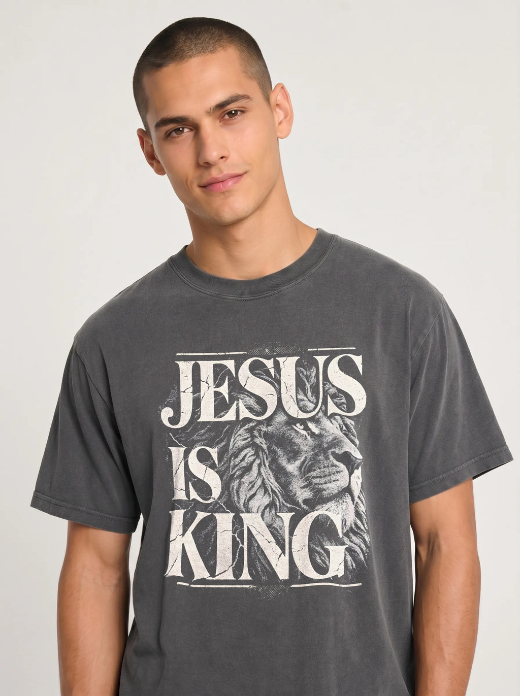 A man wearing a dark gray graphic t-shirt with a distressed white print that reads 'JESUS IS KING' alongside an illustration of a lion's head, set against a plain light gray background.