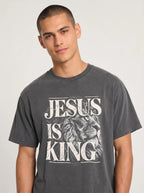 A man wearing a dark gray graphic t-shirt with a distressed white print that reads 'JESUS IS KING' alongside an illustration of a lion's head, set against a plain light gray background.