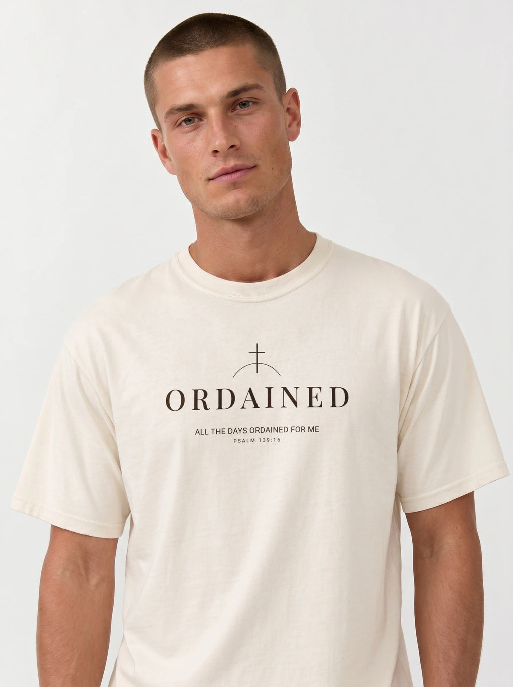 A man wearing a cream-colored crewneck t-shirt with a black graphic print that reads 'ORDAINED' and 'ALL THE DAYS ORDAINED FOR ME' below it, set against a plain light gray background.