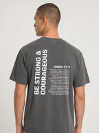 Person wearing a charcoal T-shirt with “Be strong & courageous” and Joshua 1:7–9 printed across the back.