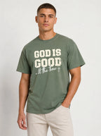 A man wearing a muted green graphic t-shirt with the text 'GOD IS GOOD all the time' in distressed white lettering, paired with light beige pants, against a plain light gray background.