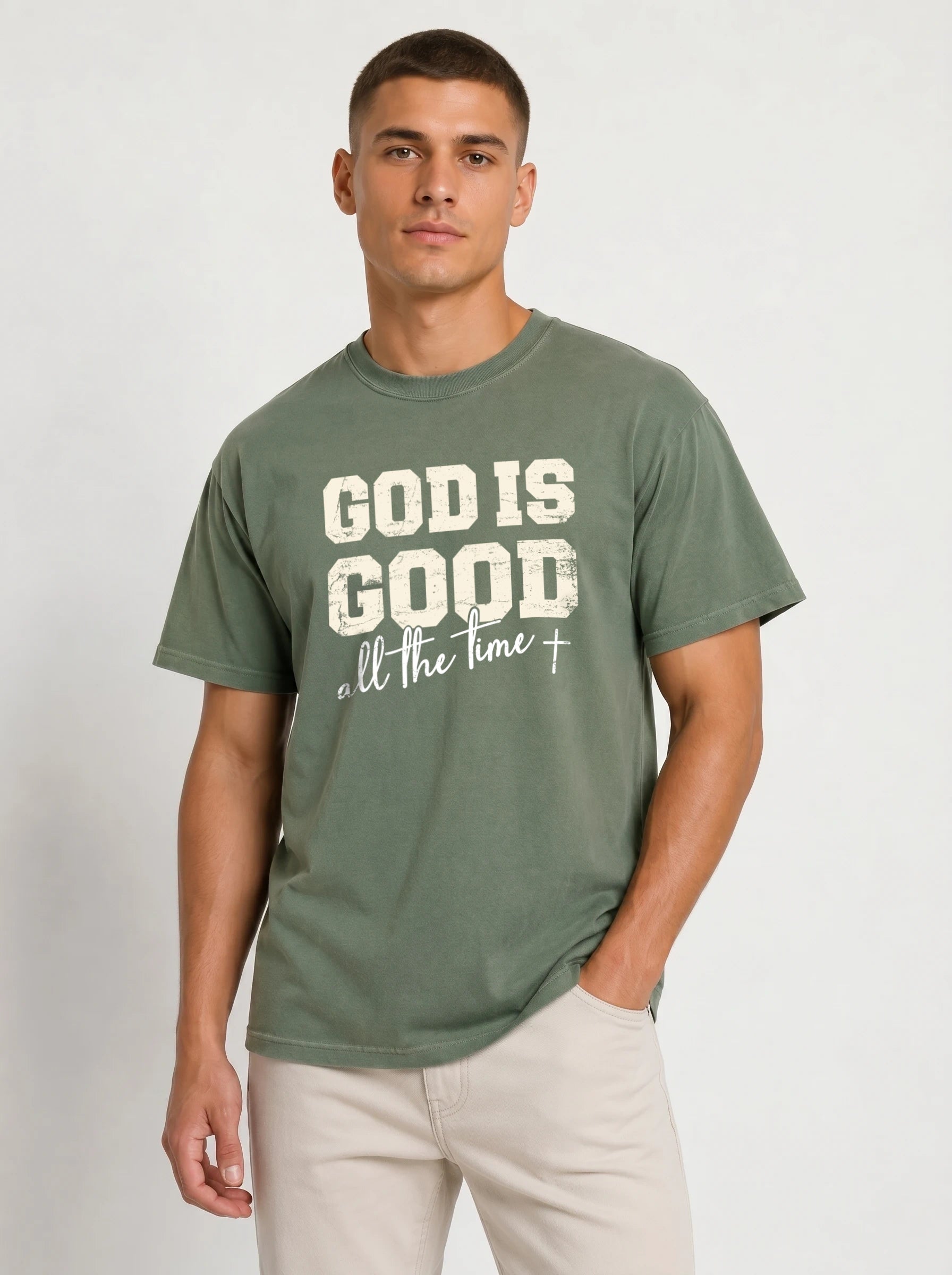 A man wearing a muted green graphic t-shirt with the text 'GOD IS GOOD all the time' in distressed white lettering, paired with light beige pants, against a plain light gray background.