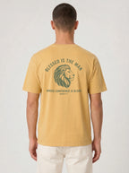 A man viewed from the back wearing a mustard yellow t-shirt with a green graphic of a lion's head and the text 'BLESSED IS THE MAN WHOSE CONFIDENCE IS IN HIM' on the back, paired with off-white pants against a light gray background.