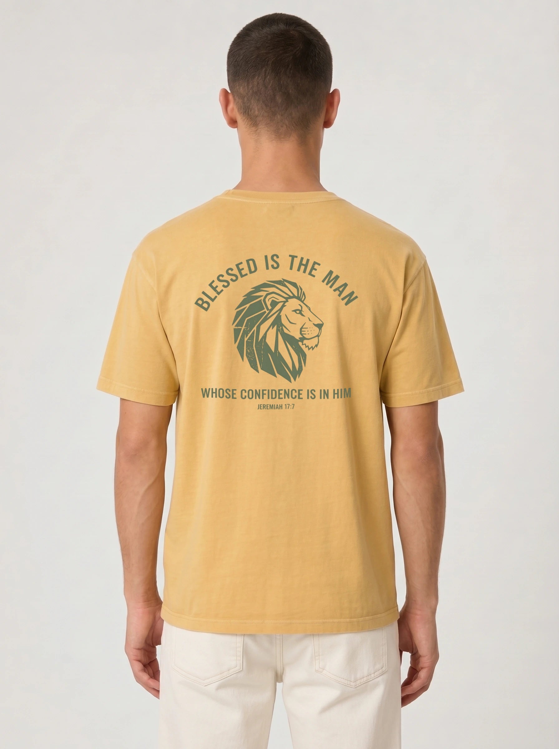A man viewed from the back wearing a mustard yellow t-shirt with a green graphic of a lion's head and the text 'BLESSED IS THE MAN WHOSE CONFIDENCE IS IN HIM' on the back, paired with off-white pants against a light gray background.
