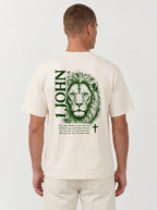 Person wearing a cream T-shirt with a green lion illustration, cross, “1 John 4:4,” and the verse text, “You, dear children, are from God and have overcome them, because the one who is in you is greater than the one who is in the world.”