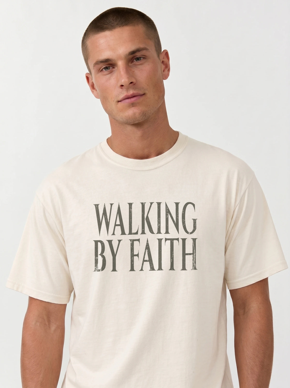 Person wearing a cream T-shirt with distressed gray text reading “Walking By Faith.”