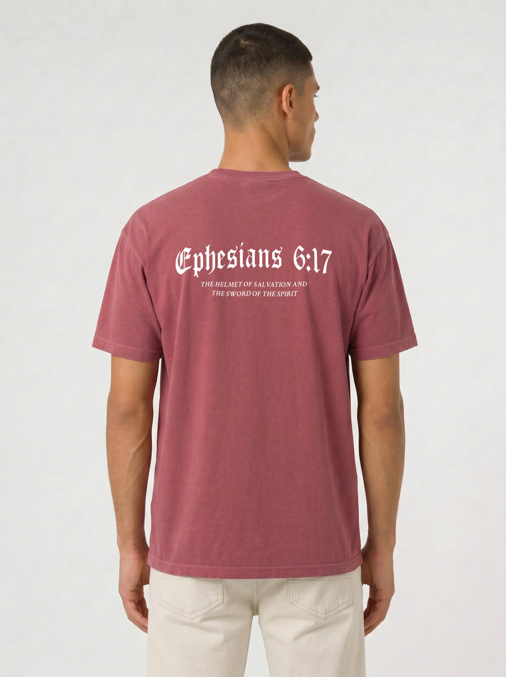 A man viewed from the back wearing a heathered dusty rose t-shirt with 'Ephesians 6:17' and smaller text underneath, paired with off-white trousers, against a light gray studio background.