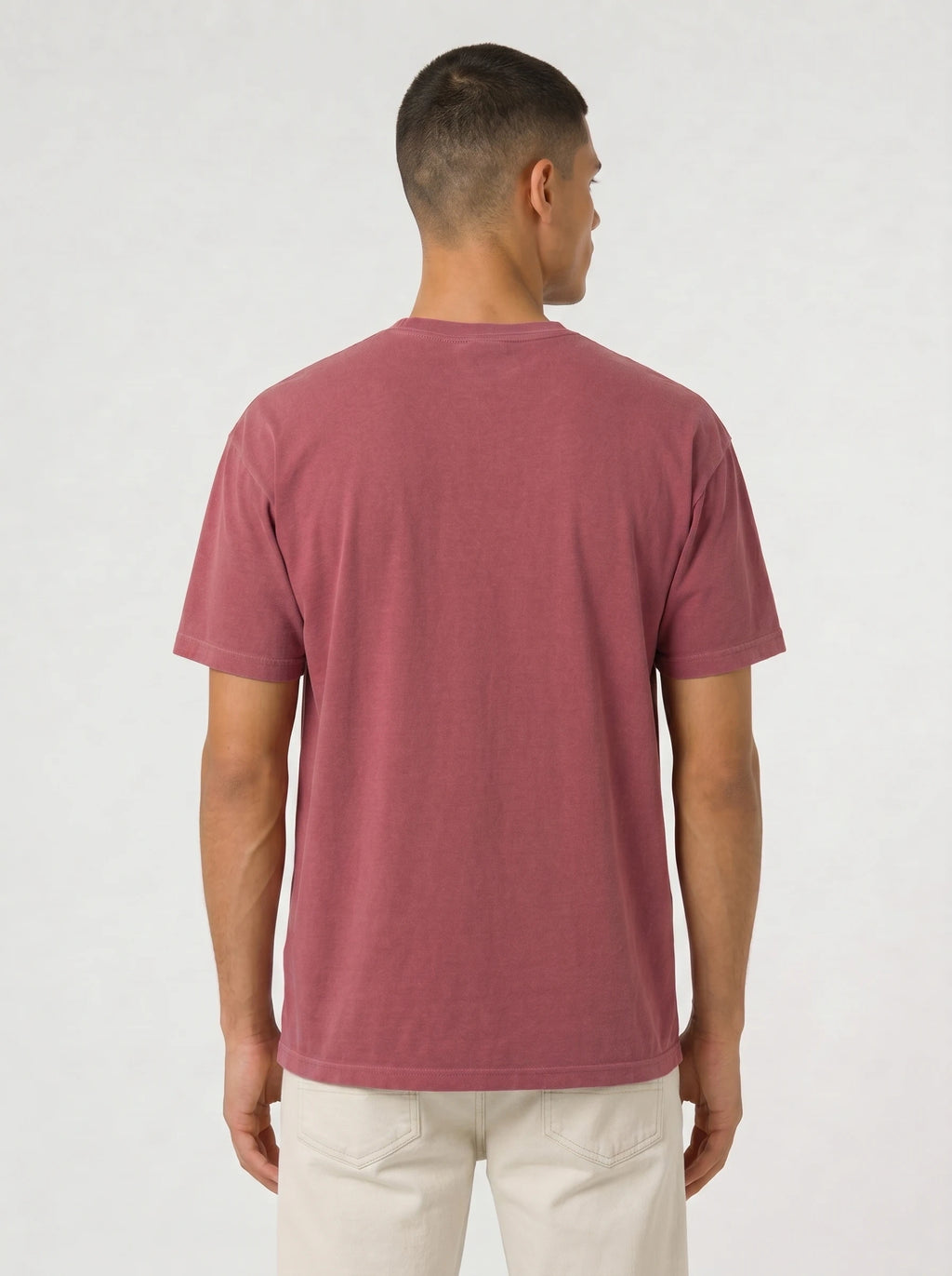 A man viewed from the back wearing a muted dusty rose short-sleeve t-shirt and off-white trousers, standing against a plain light gray studio background.