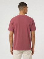 A man viewed from the back wearing a muted dusty rose short-sleeve t-shirt and off-white trousers, standing against a plain light gray studio background.