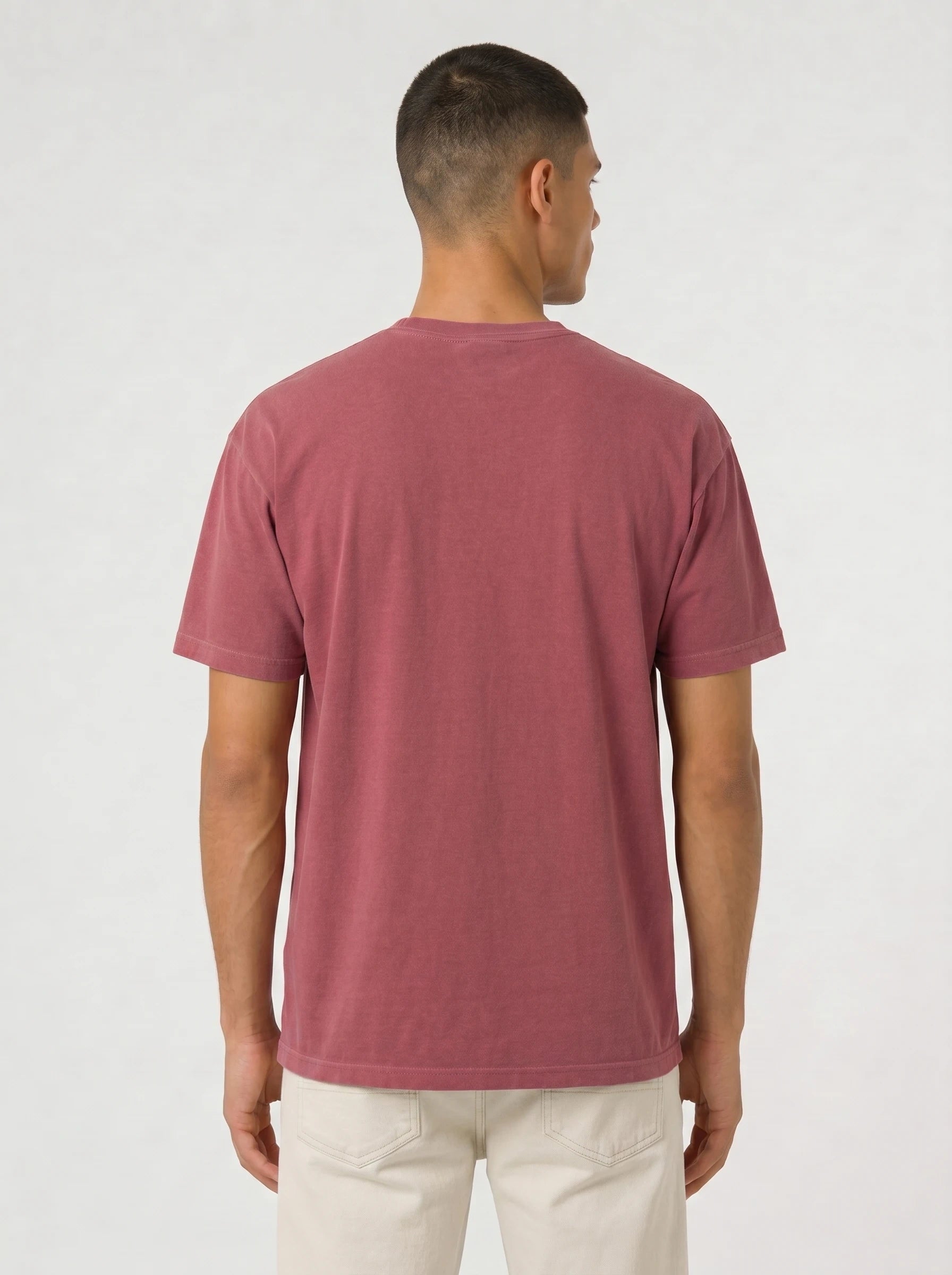 A man viewed from the back wearing a muted dusty rose short-sleeve t-shirt and off-white trousers, standing against a plain light gray studio background.