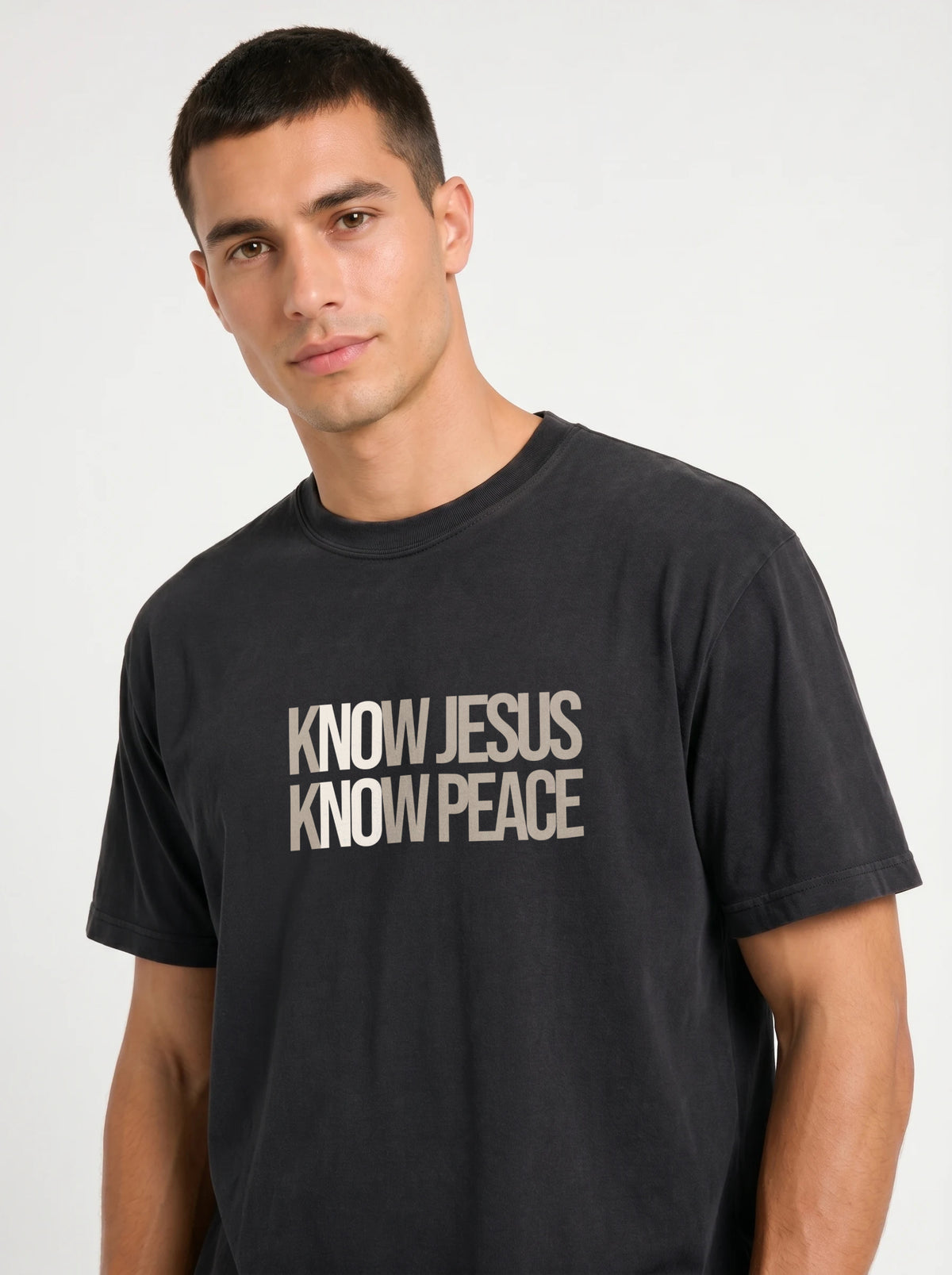A man wearing a faded black t-shirt with the text 'KNOW JESUS KNOW PEACE' printed in a distressed beige font on the chest, photographed against a light gray background.