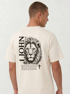 Person wearing a cream T-shirt with a black lion illustration, cross, “1 John 4:4,” and the verse text, “You, dear children, are from God and have overcome them, because the one who is in you is greater than the one who is in the world.”