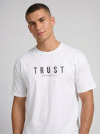 A man wearing a white crew-neck t-shirt with the 'TRUST' printed in black across the chest, set against a plain light gray background.