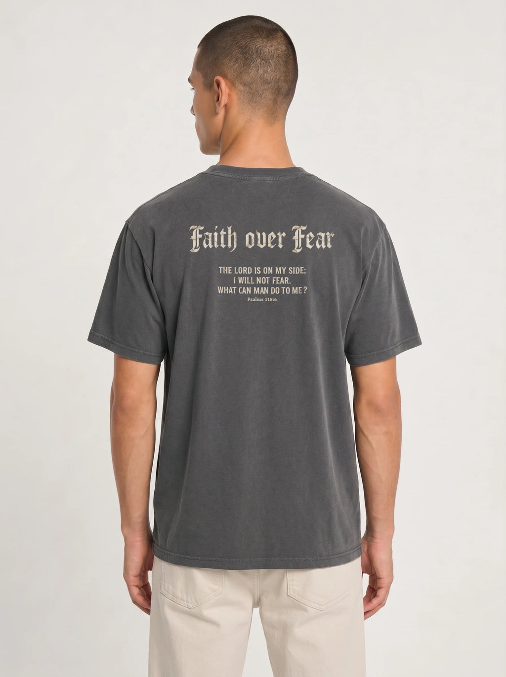 A man viewed from the back wearing a dark gray t-shirt with a sand gothic-style text graphic reading 'Faith over Fear' and a bible verse, paired with light beige pants, against a plain light gray background.