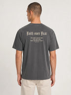 A man viewed from the back wearing a dark gray t-shirt with a sand gothic-style text graphic reading 'Faith over Fear' and a bible verse, paired with light beige pants, against a plain light gray background.