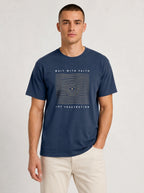A man wearing a navy blue graphic t-shirt with a wavy line design and the text 'WAIT WITH FAITH' and 'NOT FRUSTRATION' on the front, paired with light beige pants, against a light gray studio background.