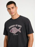 A man wearing a faded black graphic t-shirt with a white and pink fish design and the text 'FISHERS OF MEN' and 'MATTHEW 4:19'.