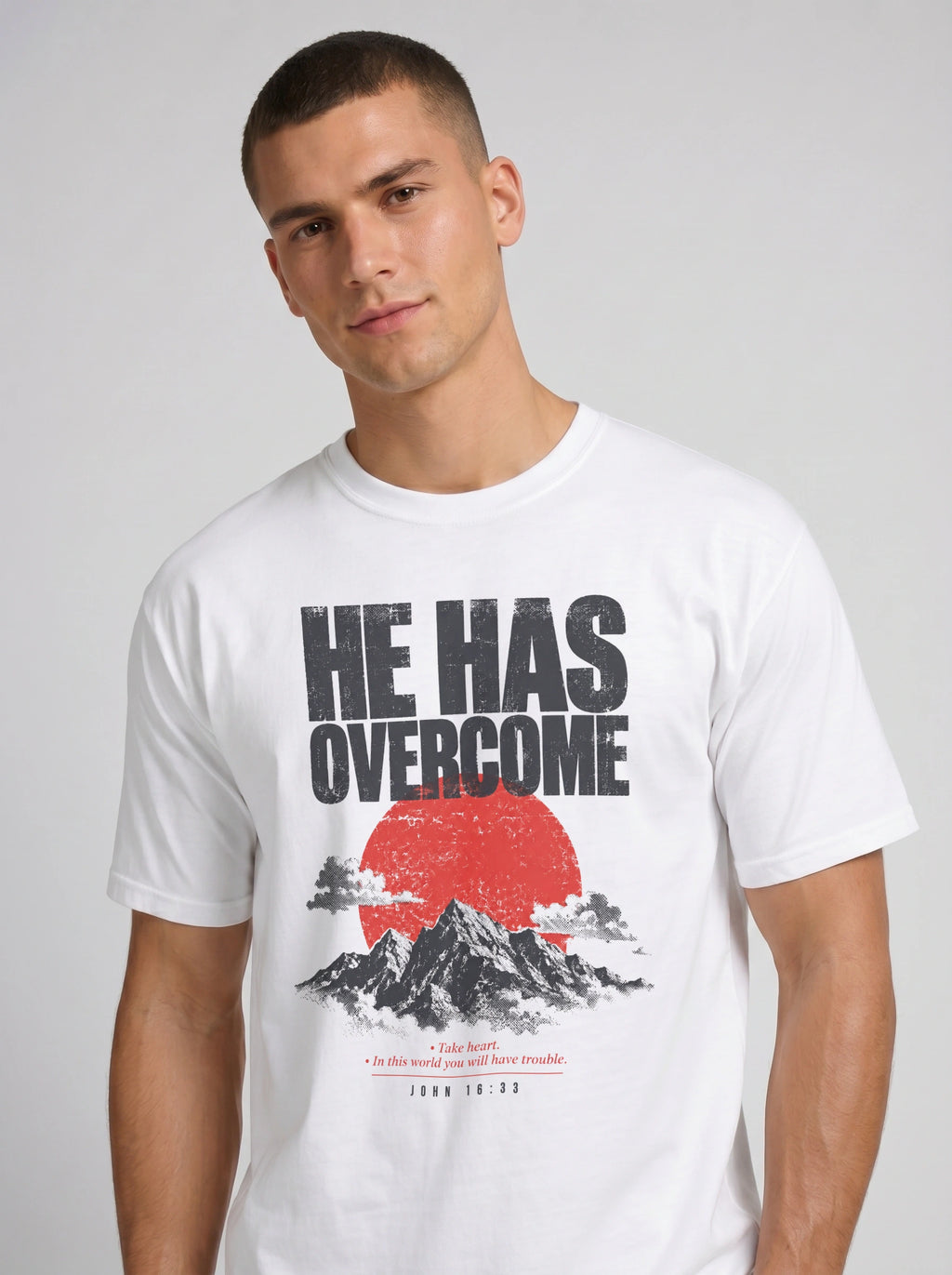 A man wearing a white crew-neck t-shirt with a graphic print that reads 'HE HAS OVERCOME' above a red sun and mountain illustration, set against a plain light gray background.