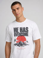 A man wearing a white crew-neck t-shirt with a graphic print that reads 'HE HAS OVERCOME' above a red sun and mountain illustration, set against a plain light gray background.