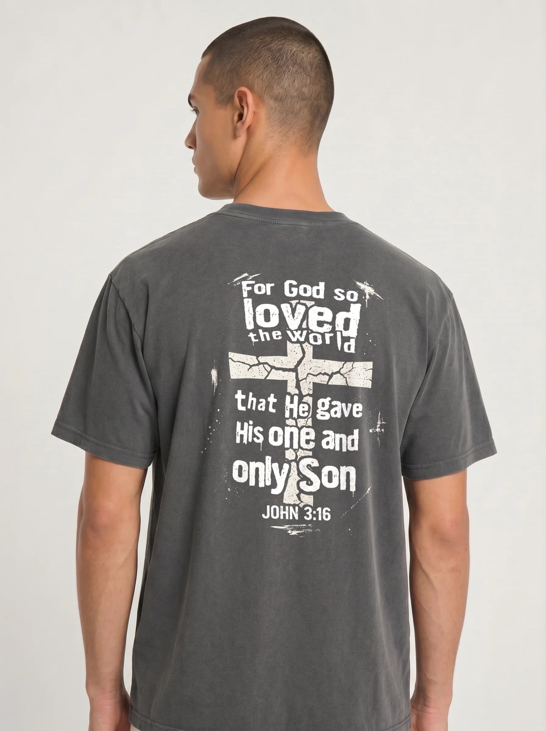 A man wearing a dark gray t-shirt, viewed from the back. The shirt features a large white distressed graphic with a cross and a Bible verse from John 3:16.