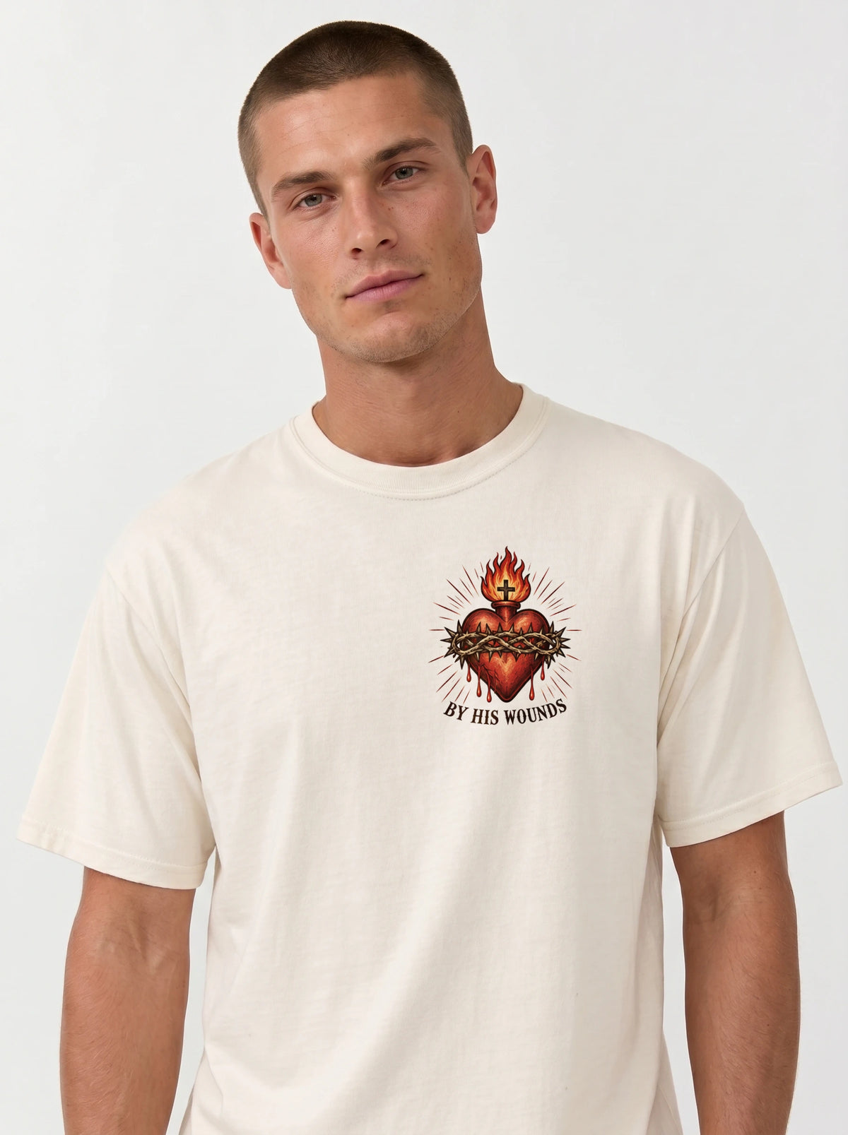 Cream T-shirt with a flaming sacred heart wrapped in a crown of thorns and the text “By His Wounds.”