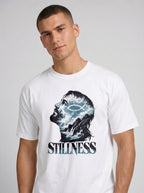 A man wearing a white crewneck t-shirt with a graphic print of a profile head filled with stormy blue and white clouds, featuring the word 'STILLNESS' in a stylized font below it.