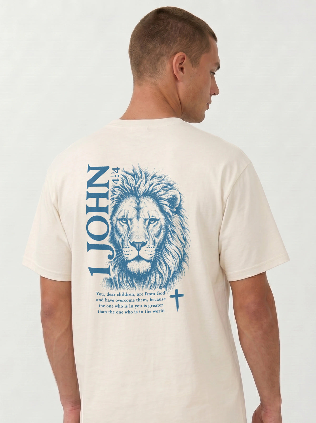 A man wearing a cream-colored garment-dyed t-shirt, viewed from the back. The shirt features a large blue lion illustration with '1 JOHN 4:4' text, a Bible verse beneath it, and a small cross symbol.