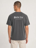 A man viewed from the back wearing a dark gray washed t-shirt with 'Psalm 23:4' printed in white Gothic lettering on the upper back, paired with light beige pants, against a light gray studio background.