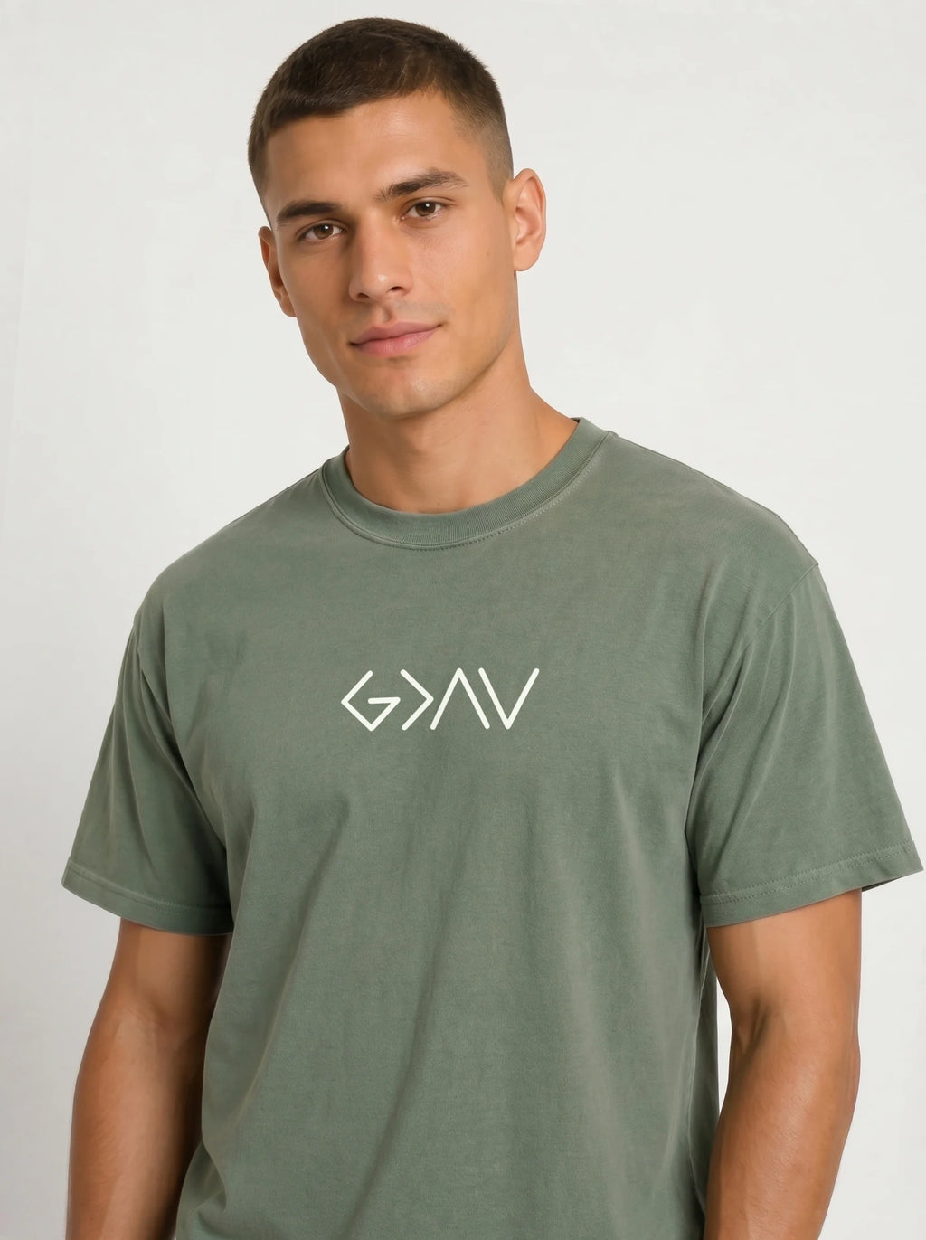 A man wearing a muted sage green crewneck t-shirt with a white stylized 'God Is Greater Than Highs and Lows' logo printed on the chest, photographed against a light gray studio background.