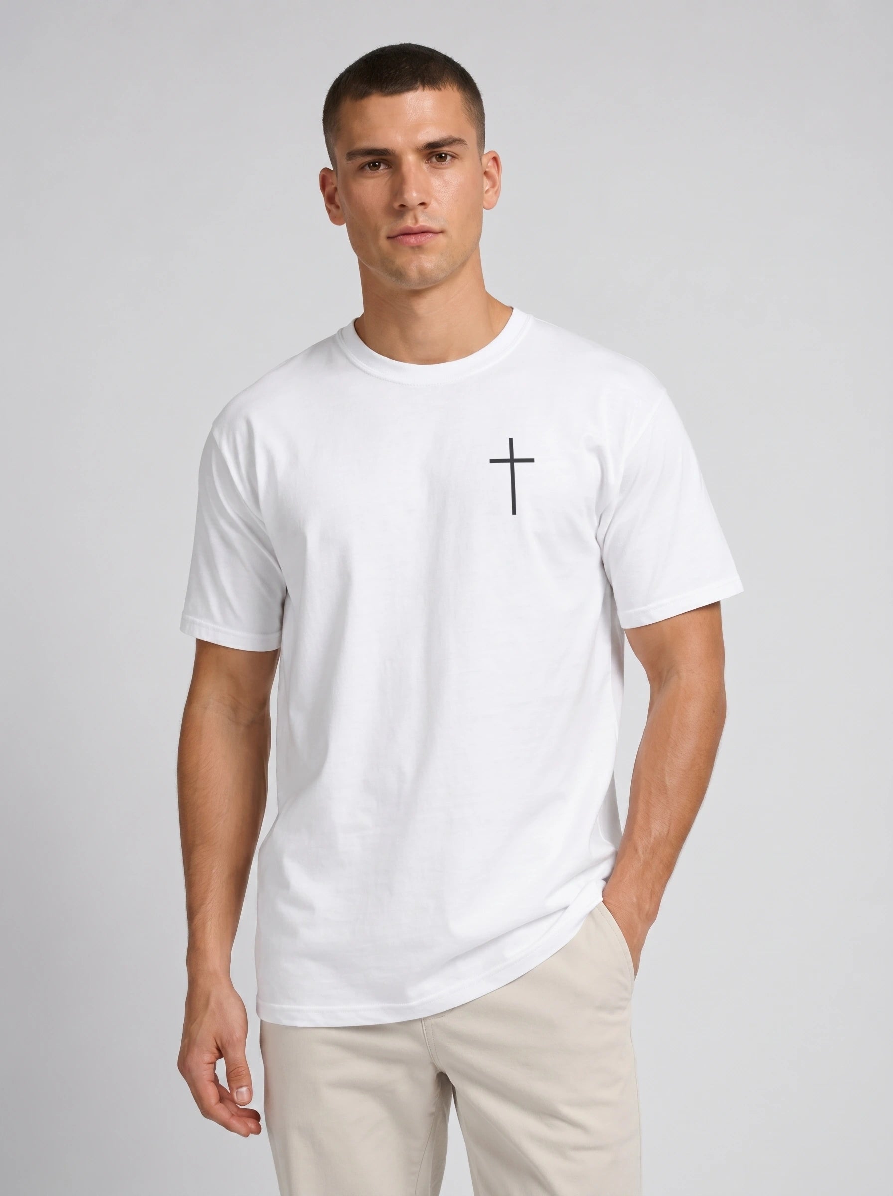 A man wearing a white short-sleeve crewneck t-shirt with a small black cross printed on the left chest, paired with beige trousers, against a light gray studio background.