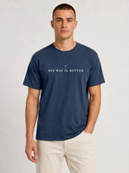 A man wearing a navy blue crewneck t-shirt with the white printed text 'HIS WAY IS BETTER' and a small cross in the center, paired with light beige pants, against a light gray studio background.