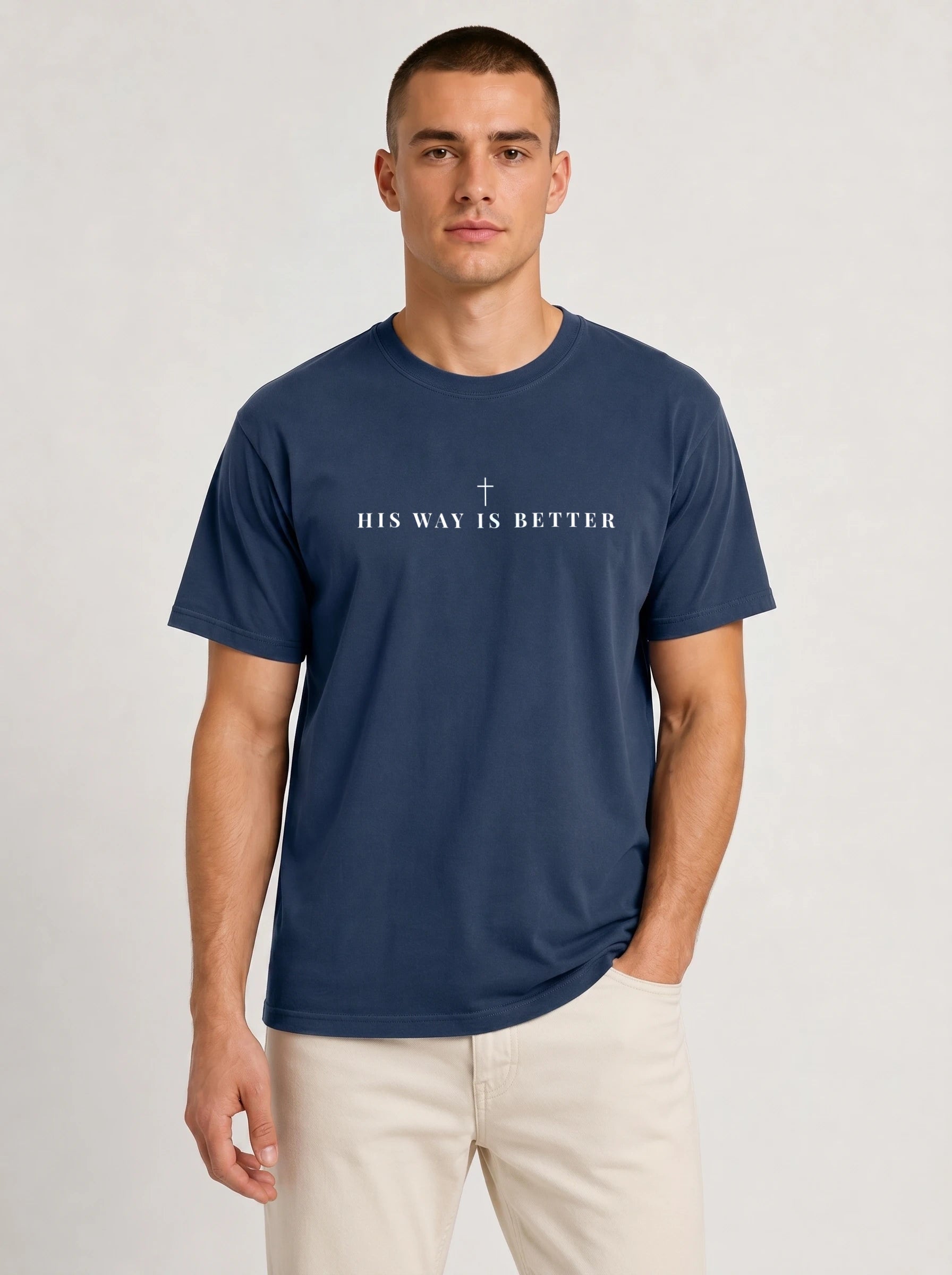 A man wearing a navy blue crewneck t-shirt with the white printed text 'HIS WAY IS BETTER' and a small cross in the center, paired with light beige pants, against a light gray studio background.